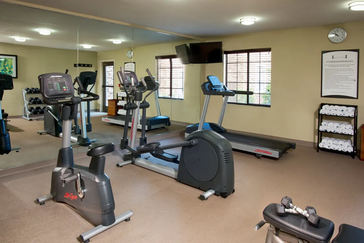 A bright, motivating interior fitness center, complete with modern exercise bikes, treadmills, dumbbells, and a mirrored wall that reflects the clean, well-organized space. The fitness rules poster on the wall adds a thoughtful hotel-style touch. The setting conveys both personal health goals and opportunities for group activity.