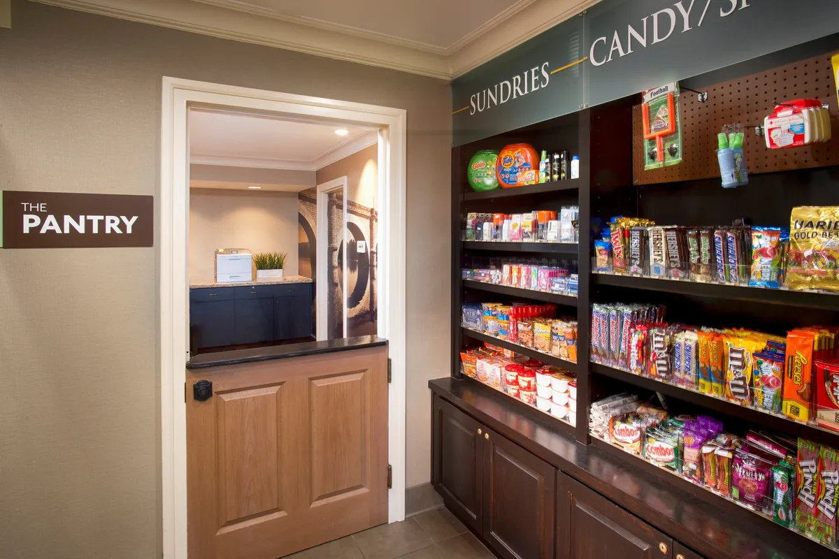 A casual, inviting pantry area with shelves stocked full of snacks and drinks. A sign reading 'THE PANTRY' and an open doorway invite guests to explore and pick up essentials or treats at their convenience.