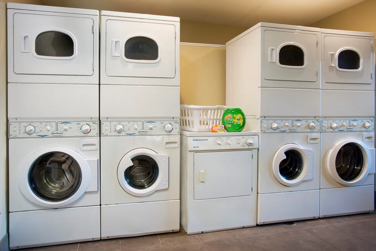 A tidy laundry facility with stacked washer and dryer units, a laundry basket, and detergent, set against a simple, efficient backdrop. The scene highlights the convenience of in-building laundry for guests staying longer or seeking independence.