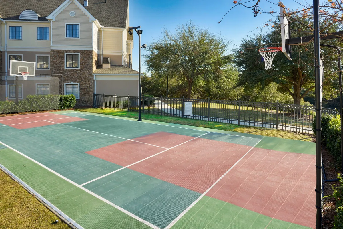 An energetic exterior shot of a basketball court nestled within lush greenery, featuring a prominent hoop and open space, inviting friendly games and playful competition. The setting sun creates a warm, golden glow over the scene.