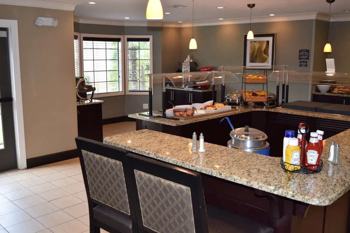 A cozy breakfast area with granite countertops displaying a spread of food and beverages. Large windows allow natural light to fill the room, where families can gather to enjoy a complimentary hot breakfast, socialize, and prepare for their day of fun.