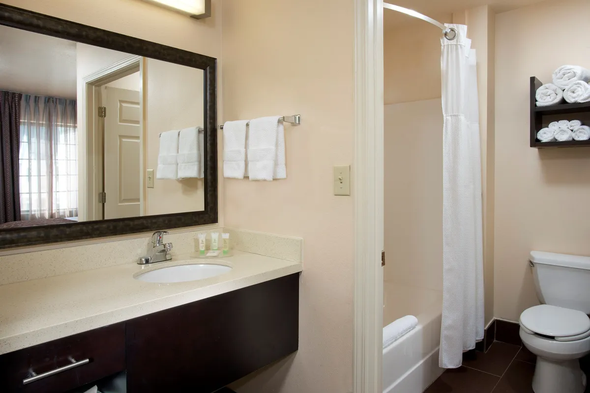 This image depicts a bright, clean bathroom with a modern sink featuring neatly arranged toiletries, fluffy towels, a large mirror, and a visible shower area. The overall atmosphere is calm and inviting, with a focus on hotel-level cleanliness and comfort.