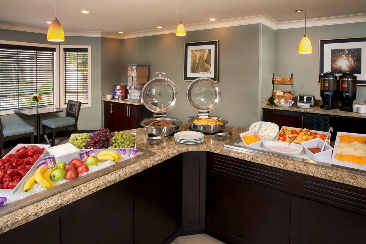 A welcoming breakfast buffet area with a spread of morning favorites and ample seating. The space is lively, with guests socializing and enjoying their meals surrounded by vibrant decor and decorative artwork. The buffet tables are well-stocked, underscoring the appeal of the complimentary on-site breakfast.