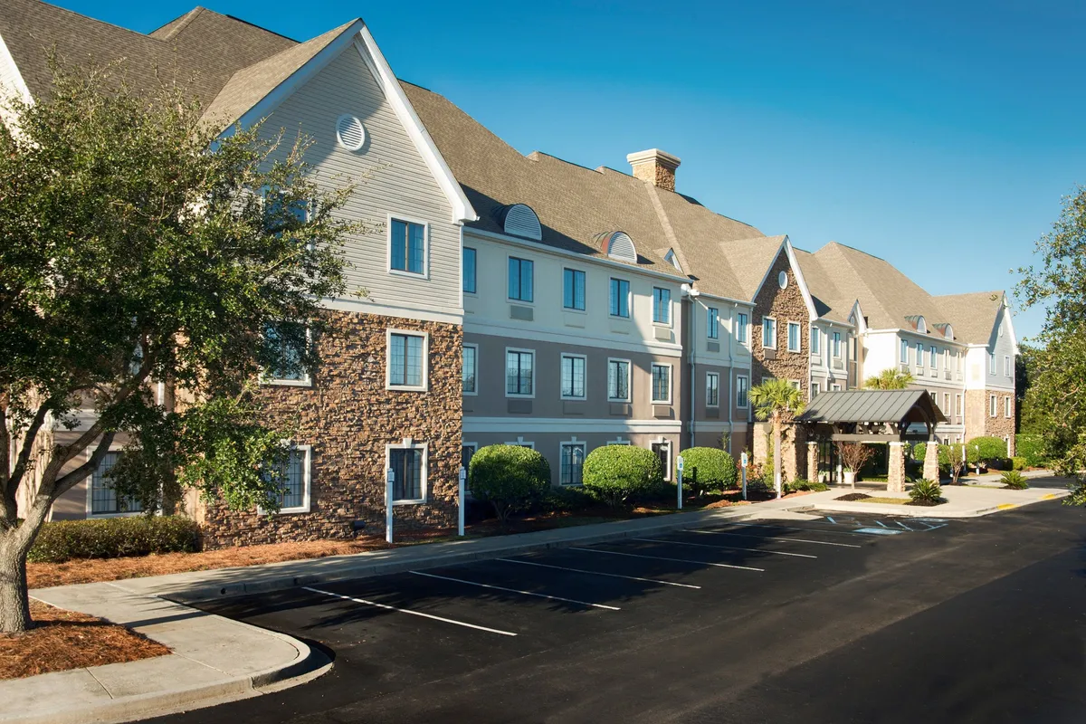The main entrance of the property, featuring a welcoming awning, attractive stone facade, neatly landscaped bushes, and bright blue windows. The scene suggests easy accessibility, arrival excitement, and proximity to local attractions, making it ideal for families starting their adventures.