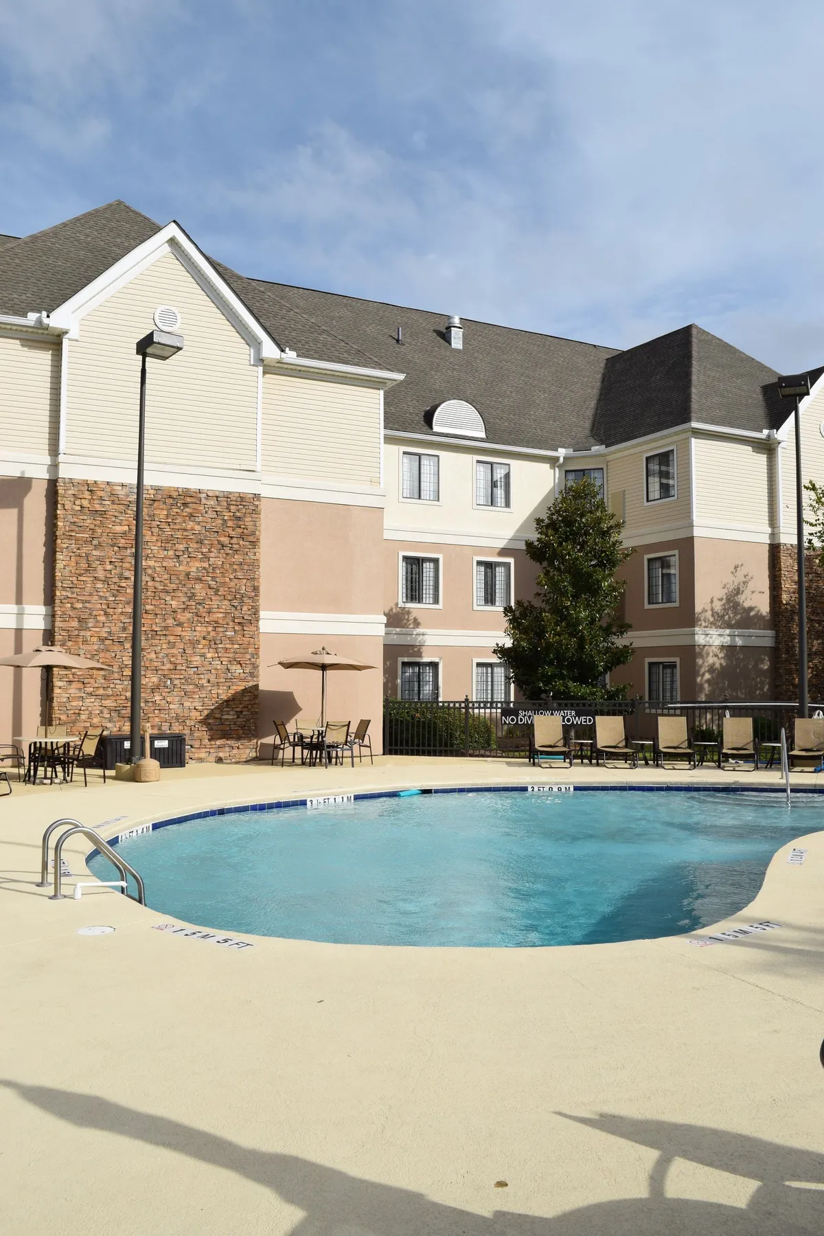 A vibrant exterior scene featuring a sparkling swimming pool surrounded by an inviting outdoor seating area, the hotel’s façade rising above, and well-maintained landscaping. Guests are seen swimming, sunbathing, and relaxing in groups, capturing a lively, social atmosphere.