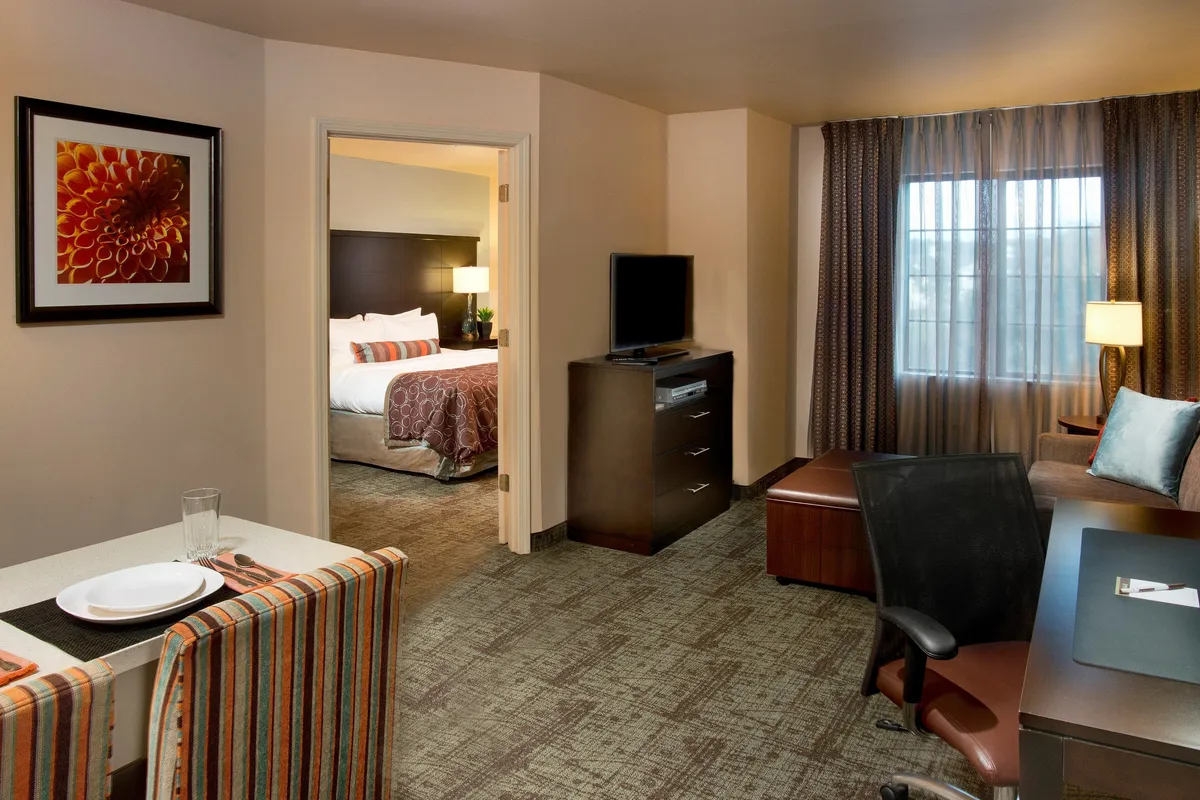 The image shows a bright, inviting condo interior featuring a dining table set with plates and glasses, perfect for gathering over meals. Beyond the dining area, the bedroom doorway is visible, and there is a cozy seating area with an armchair and a television, creating a comfortable space for both relaxation and productivity.