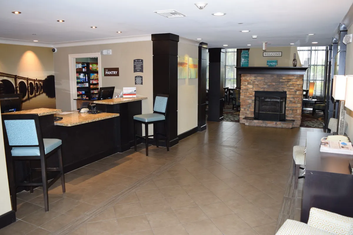 A welcoming hotel lobby featuring a check-in counter, cozy fireplace, pantry area, and comfortable seating. The space is well-lit and inviting, designed for guests to relax after a day out or while waiting for check-in.