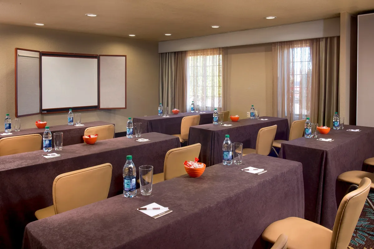 A well-lit interior space featuring tables arranged in a classroom style with a presentation board at the front. Large windows let in plenty of natural light, creating a welcoming and productive environment suitable for meetings, workshops, or working remotely.