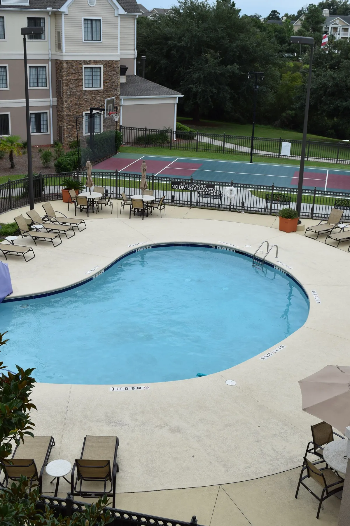 This exterior shot displays the inviting swimming pool surrounded by lounge chairs, with a clear view of a tennis court adjacent to the pool area. The image captures people enjoying a swim and others preparing for a tennis match, with a leisurely mood and hints of sunset lighting. The spacious pool deck encourages relaxation while the adjacent sports amenities foster active fun.