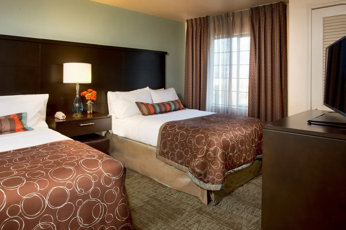 A bright and comfortable suite space with two beds, a nightstand with a lamp, a window dressed with curtains, and a television positioned for easy viewing. The room feels airy, personalized, and conducive to relaxing or watching TV together.