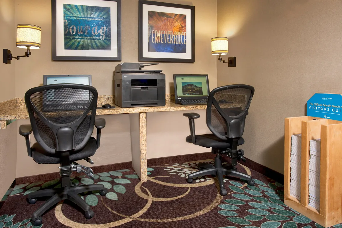 A contemporary business center interior featuring computer workstations, a printer, decorative artwork, and a rack of visitor guides. The ambiance is productive yet welcoming, ideal for business travelers or guests needing to check emails or plan outings.